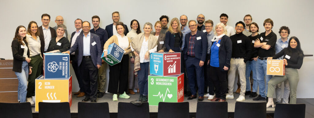Personen, Workshop-Leader, Helfende des Symposiums, Keyspeaker, usw., posieren für ein Gruppenfoto auf einer kleinen Bühne. Dazwischen auf der Bühne und in den Händen sind manchmal unterschiedlich grosse Würfel mit SDGs darauf verteilt.